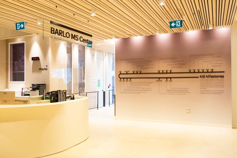 The bright, wood-panelled lobby of the BARLO MS Centre, with a curved reception desk and an 'MS Milestones' timeline wall.
