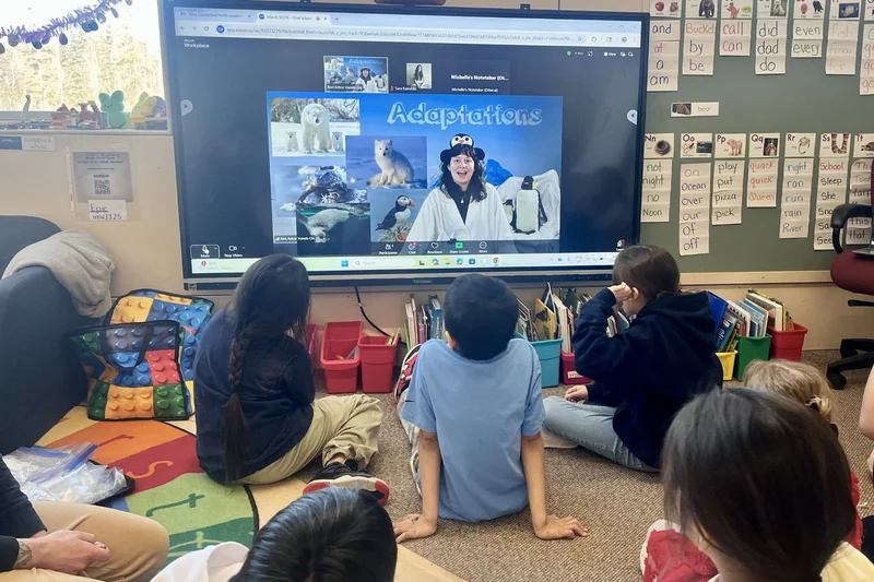 Young students sit together on a classroom floor, watching a virtual lesson on animal adaptations presented on a large screen.