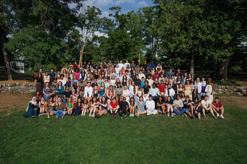 A large group of scholarship recipients gathered outdoors on a sunlit lawn, standing and seated together in front of tall trees.